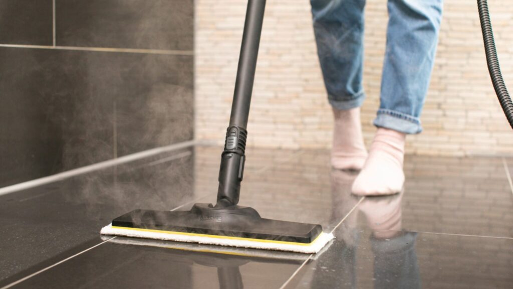Steam mop emitting vapor as it cleans a glossy black tile floor, person in rolled jeans and socks standing behind.