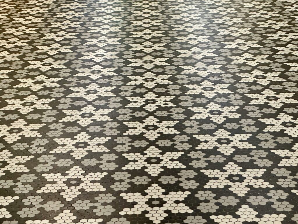 A geometric tile floor pattern featuring a repeating design of white and gray hexagonal tiles arranged in floral star shapes on a dark background.