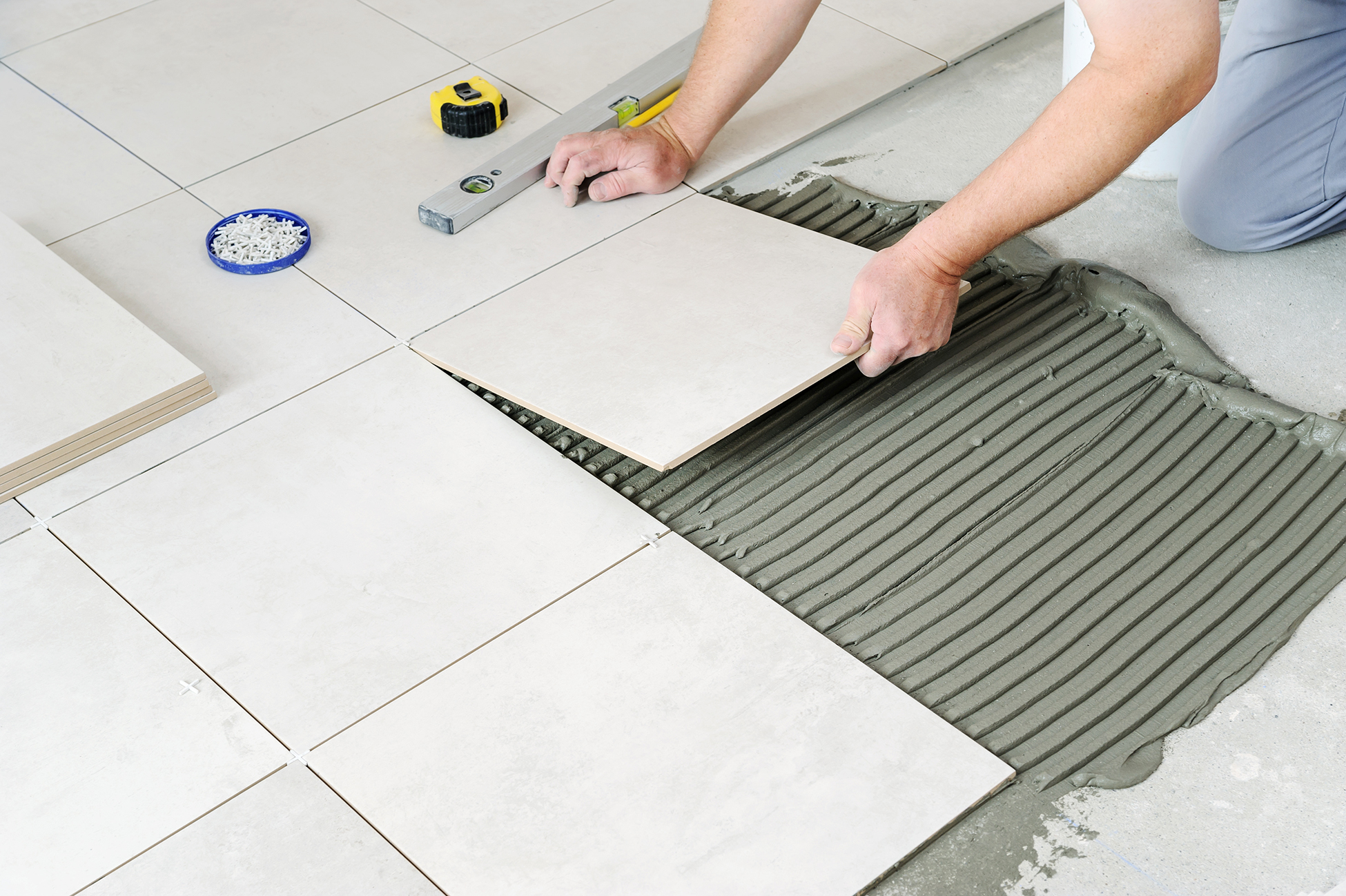 Close-up of a beginner tile installer installing large square ceramic tiles on a floor using tile adhesive and a notched trowel, with tile spacers, a level, and measuring tape nearby