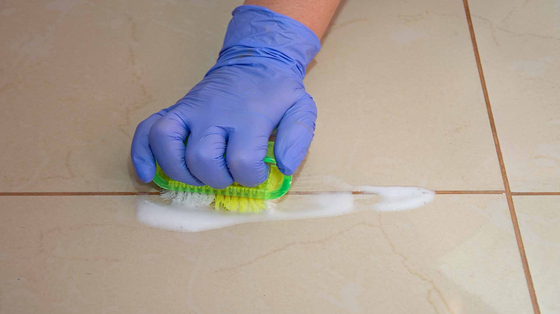 Hand in purple latex glove scrubbing beige tile grout with a green brush and foamy cleaner.