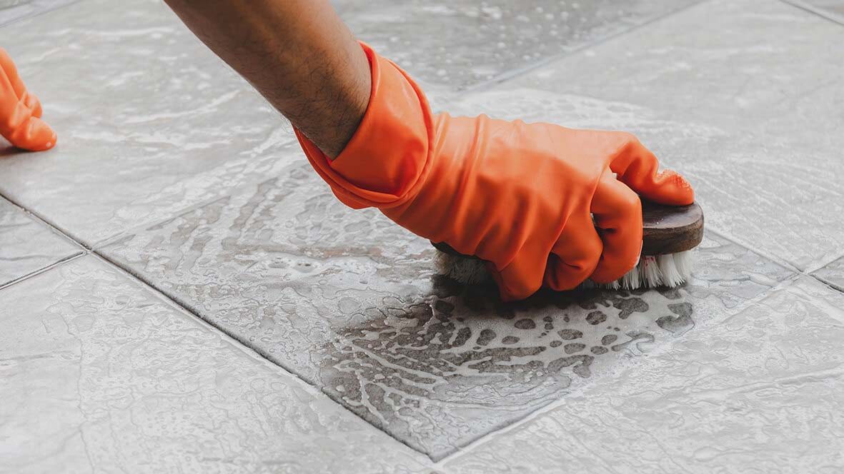 Hand in orange rubber glove scrubbing soapy tile grout with a stiff brush.