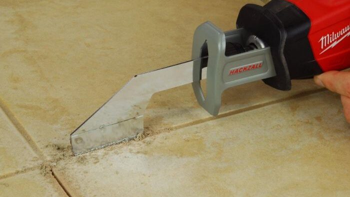 Reciprocating saw with grout‑removal blade cutting dusty grout from beige floor tiles.